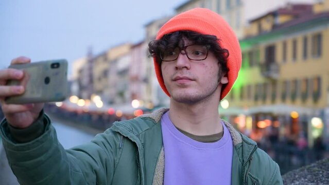 A cute young Caucasian man wearing glasses and an orange cap is using his mobile phone while resting on one of the bridges that crosses Milan's canals. He poses for a selfie.