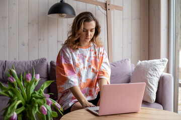 Portrait of woman typing on modern pink laptop and sitting on soft sofa in casual clothes near table with vase of fading tulips. Living room in elegant design with aesthetic decor, free copy space