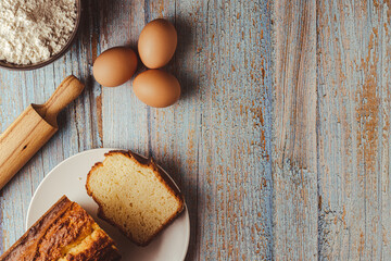 Aerial photo of a weathered wooden work table with eggs, a whisk, a rolling pin, flour and a piece of sponge cake on a plate.