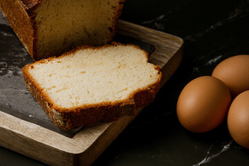 close up photo of sponge cake on wooden and stone board with fresh eggs on black marble background