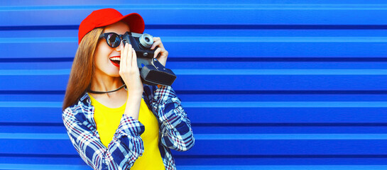 Portrait of happy young woman photographer taking picture on film camera wearing red baseball cap on blue background, banner blank copy space for advertising text