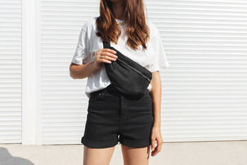 Woman, wearing white t-shirt, black shorts and fanny pack or waist pack, standing outdoor near white wall. Details of stylish trendy basic minimalistic casual outfit. Street fashion. No face.