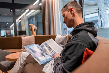 St. Anton am Arlberg. March 10, 2022. Young man reading magazine while sitting on sofa in hotel lobby, Young man reading magazine while relaxing on sofa in chalet