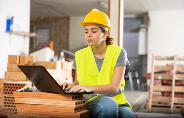 Professional young female civil engineer checking house renovation plan and blueprints on laptop at construction site indoors