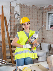 Portrait of young female builder wearing protective gear posing with handheld demolition hammer at indoor construction site