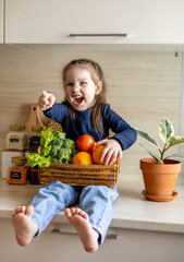 Fototapeta premium Baby sits on the kitchen table and holds a wicker basket of fruit in her hands.