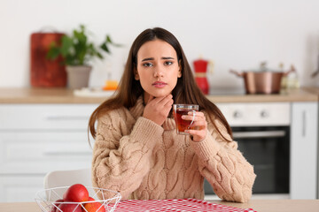 Young woman with cup of tea suffering from sore throat at table in kitchen