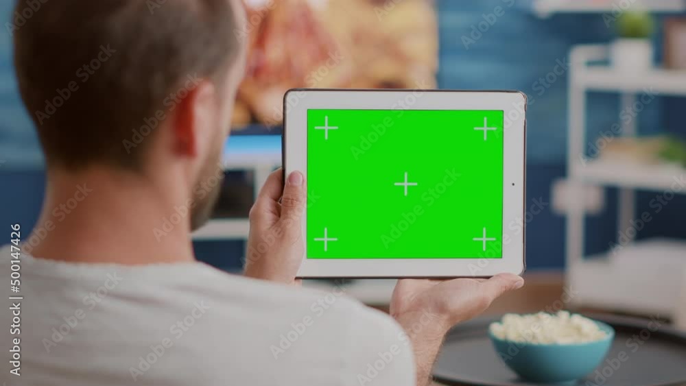 Poster closeup of man holding digital tablet with green screen watching webinar sitting on sofa in front of