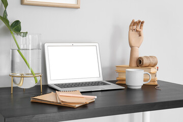 Laptop with plant leaf in vase, wooden hand and cup on table near white wall