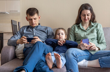Family sit on the couch and hang out on their phones. Mom dad and daughter spend time with gadgets.