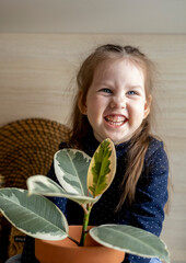 Toddler girl sits on the kitchen table sits and holds a clay pot with ficus in her hands.