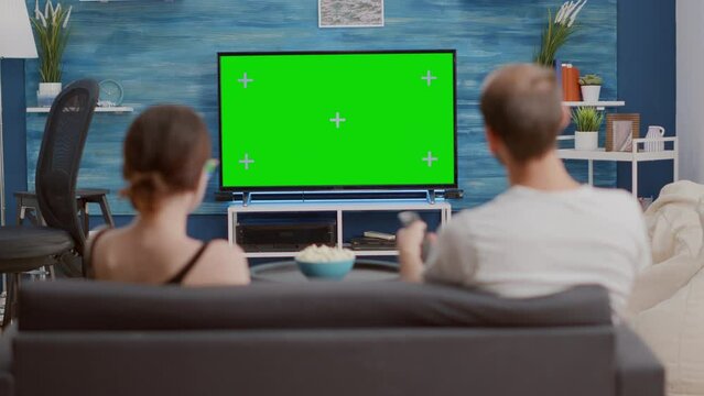 Couple sitting on couch watching television on green screen flat tv talking about favourite show and zapping in modern living room. Man and woman having popcorn enjoying movie on chroma key display.