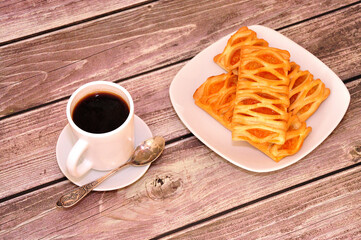 Puff pastries with apricot jam on a plate and a cup of hot black coffee on a wooden table.