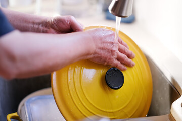 Man hands washing cleaning beautiful yellow cast iron cooking pot lid and dishes after dinner at...