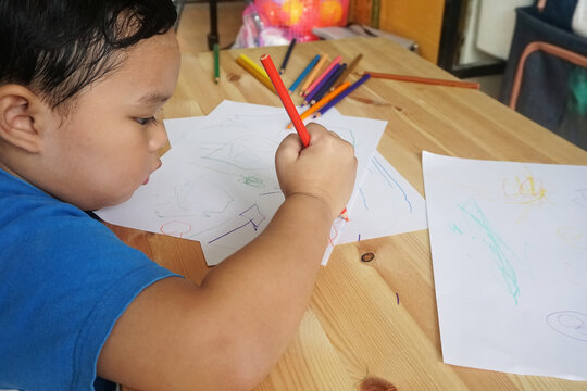 Cute Asian boy drawing with color pencils and white paper on wooden table