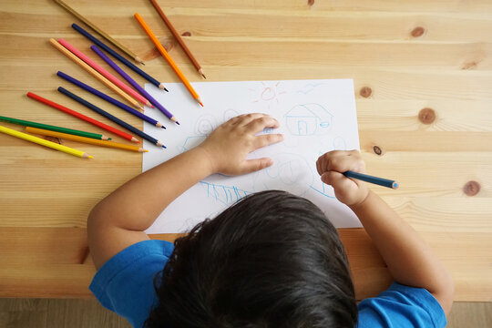 Cute Asian boy drawing happy family and home. with color pencils and white paper on wooden table, top view