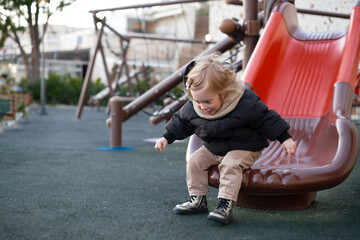 Fototapeta na wymiar Cute adorable pretty caucasian blonde baby girl sliding,having fun.Happy, warm clothing child,kid,toddler, infant of 1-2 year old in playground .Lifestyle outdoors, copy space for text