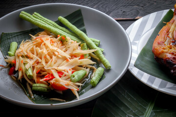 Thai food dish both in Thailand and Asia, "Papaya Salad" or as we call it "Somtum" is complemented with grilled chicken and sticky rice with fresh stir-fries. Served on the black wooden table.