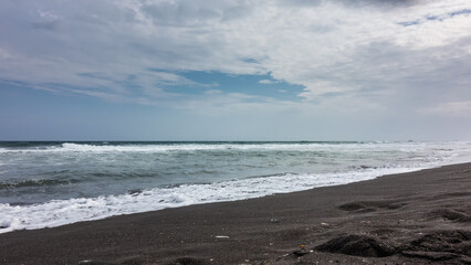 Long waves roll and foam on the beach with black volcanic sand. Clouds in the blue sky. Kamchatka. Pacific ocean. Khalaktyrsky beach