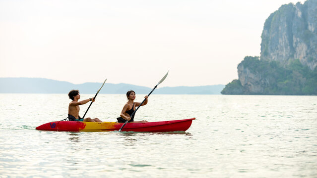 Young Asian Man And Woman Kayaking Together In The Sea At Tropical Lagoon Island At Summer Sunset. Male And Female Friends Enjoy Outdoor Activity Lifestyle And Water Sports On Beach Holiday Vacation