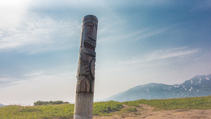 There is an old weathered wooden idol in the valley. The totem in the form of a pillar is decorated with carvings. Mountains against a background of blue sky and clouds. Kamchatka. Vilyuchinsky pass.