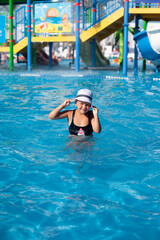 Naklejka premium Child in white panama hat is standing in water. Small happy tanned girl in black swimsuit with watermelon pattern in sunglasses poses in pool and enjoys summer vacation in water park. 