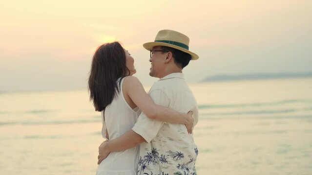 4K Happy family Asian couple holding hands and dancing together on tropical beach at summer sunset. Husband and wife relax and enjoy outdoor lifestyle romantic holiday travel beach vacation at the sea