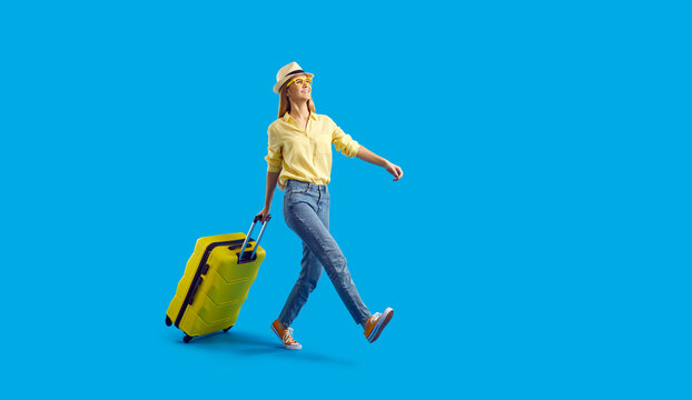 Funny cheerful tourist girl going on holiday trip. Happy young woman with yellow suitcase walking somewhere against solid blue colour studio background. Traveling concept