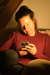 Happy millennial teen girl checking social media holding smartphone at home. Smiling young latin woman using mobile phone app playing game, shopping online, ordering delivery relax on sofa. at night.