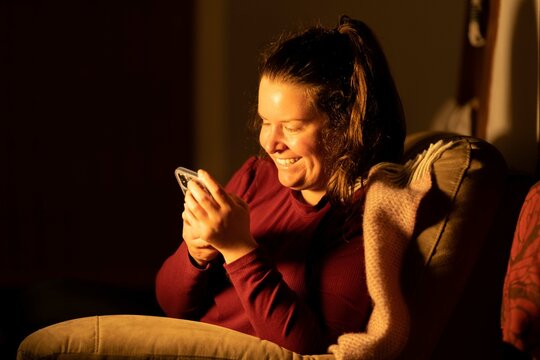 Happy Millennial Teen Girl Checking Social Media Holding Smartphone At Home. Smiling Young Latin Woman Using Mobile Phone App Playing Game, Shopping Online, Ordering Delivery Relax On Sofa. At Night.