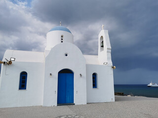 The Church of St. Nicholas the Wonderworker is white with a blue door against the backdrop of the Mediterranean Sea with a white ship and a dramatic sky.