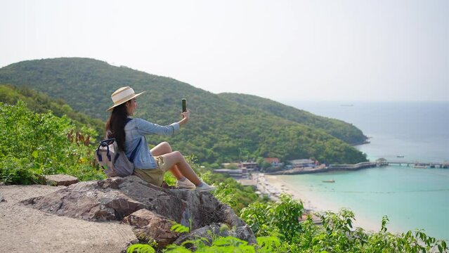 4K Young Asian woman traveler using mobile phone taking selfie while solo travel on tropical island mountain in summer sunny day. Cheerful female enjoy outdoor lifestyle in holiday beach vacation trip