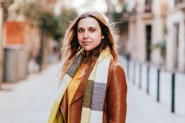 Obraz premium Charming female in leather jacket and scarf looking at camera while standing on sidewalk in city with buildings on blurred background