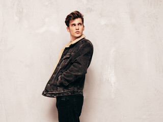 Portrait of handsome confident stylish hipster lambersexual model.Man dressed in black jacket and jeans. Fashion male posing in studio near grey wall