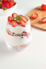 Glass with granola, yogurt and fresh strawberries on white table. Healthy breakfast bowl concept.