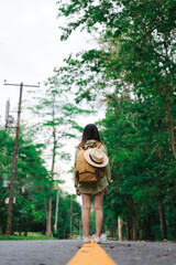Fototapeta premium Back view on a female hiker walking down the asphalt road in the woodland. Tourist woman with a backpack standing in the middle of the highway near the fir forest.