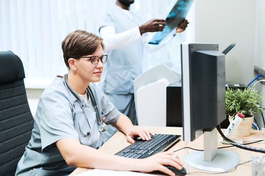 Content female doctor with stethoscope around neck working with computer in office while her colleague analyzing x-ray image