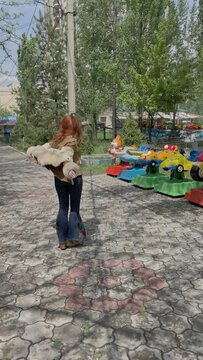 Joyful red-haired woman is spinning around herself on alley in amusement park. Children's rides and spring trees on background. Funny girl in boho fur coat and retro jeans.