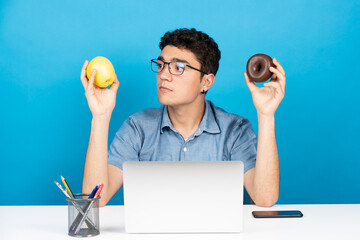 Hispanic man looking at apple and holding a chocolate donut in the other hand isolated on blue...