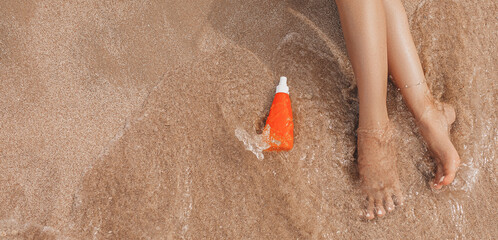 Close-up of young female bare feet with sunscreen cream on beach with a sea wave. Woman uses moisturizer, relaxing, enjoying time of summer vacation. Legs of girl on the seaside with sandy background