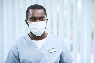 Portrait of young African American nurse in facial mask and scrubs with badge working in clinic