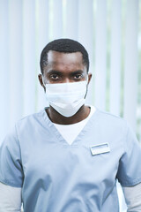 Portrait of young black medical employee in surgical mask and uniform with badge on chest standing in hospital