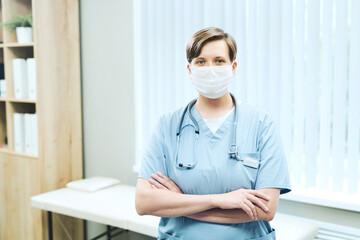 Portrait of fenale medical intern in facial mask standing with crossed arms against examination couch in hospital