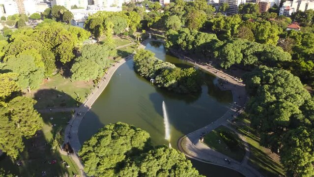 Argentine people relaxing at Centenario Park lake with Buenos Aires cityscape in background. Aerial tilt down