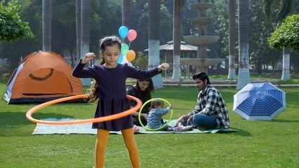 A small young Indian girl playing with a hula hoop - an outdoor sport a childhood game Indian family family picnic in a park. An Indian husband-wife spending time with their kids in the public p... - Powered by Adobe