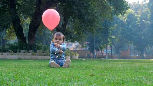 A beautiful baby girl enjoys playing with a pink colored balloon attached to a string - a fun activity  a happy childhood. An adorable infant sitting alone on the green grass in a public park - lei...
