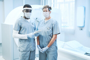 Portrait of multi-ethnic MRI specialists in scrubs and masks standing with tablet against ultra sound machine