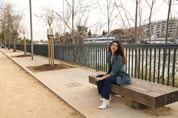 Young and beautiful woman, brunette, with curly hair wearing a jacket, jeans and white boots, sitting on a wooden bench with a thoughtful look. Concept beauty, fashion, trend, model, thoughts, autumn.