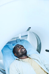 Serious young African-American patient with stubble lying on couchette of MRI machine while undergoing procedure - obrazy, fototapety, plakaty
