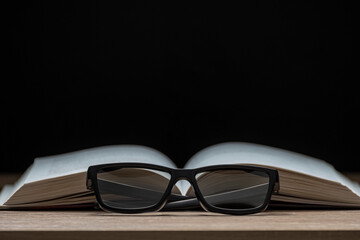 Glasses for vision and an open book lie on a table on a black background. Reading Glasses. Classic...